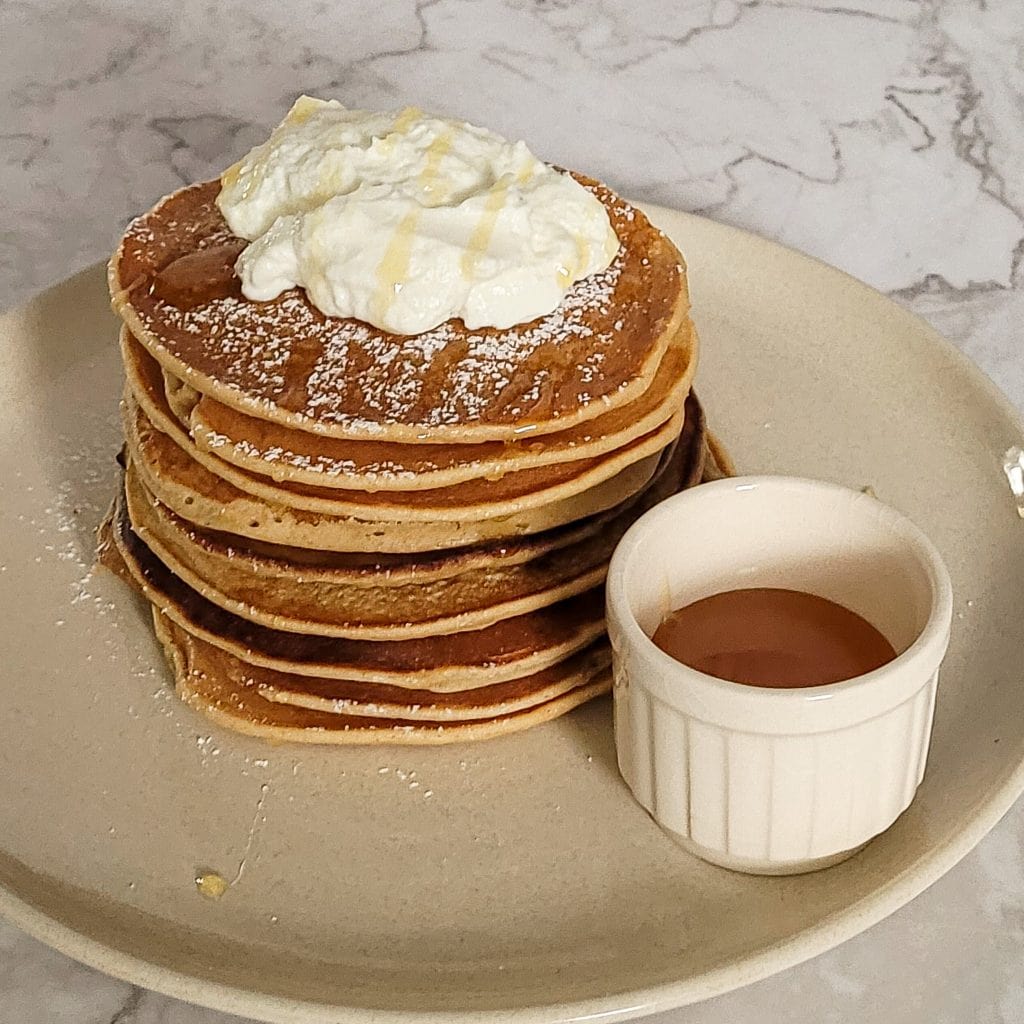 Stack of whole wheat banana pancakes with whipped cream and syrup on a plate.
