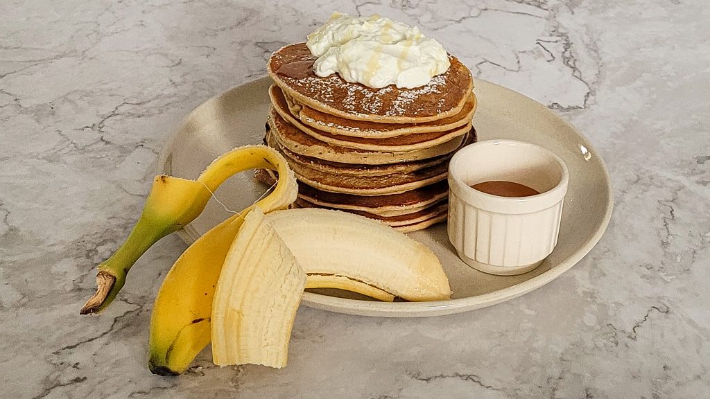 Stack of healthy banana pancakes with whipped cream and fresh bananas.