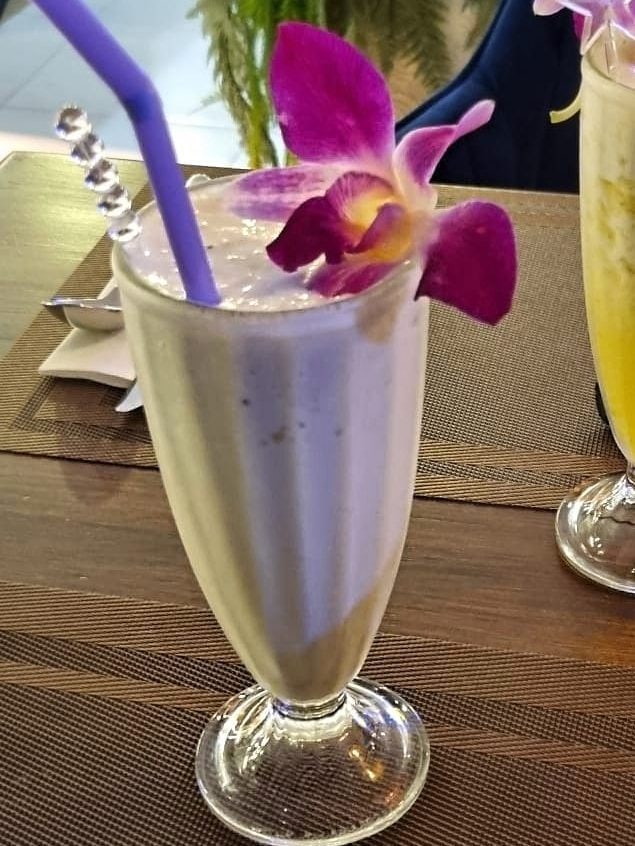 Creamy banana milkshake in a tall glass with purple straw and orchid decoration on a café table