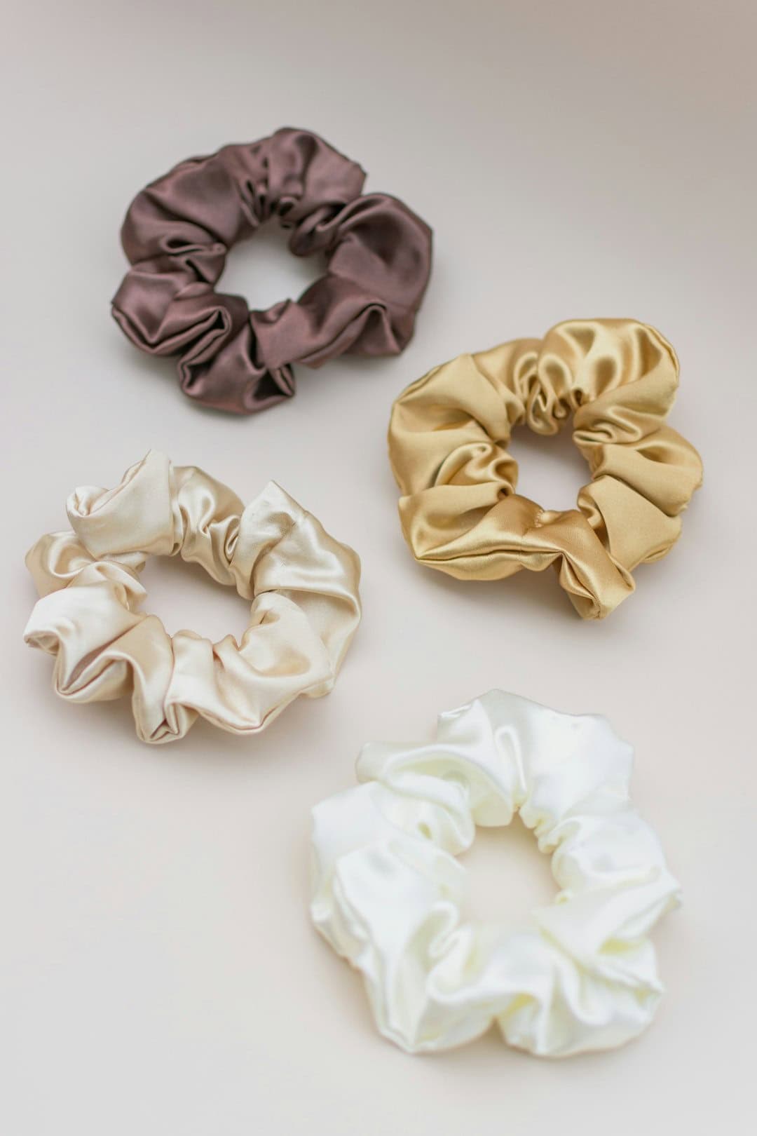 Four satin scrunchies in brown, beige, cream, and white on a neutral background.