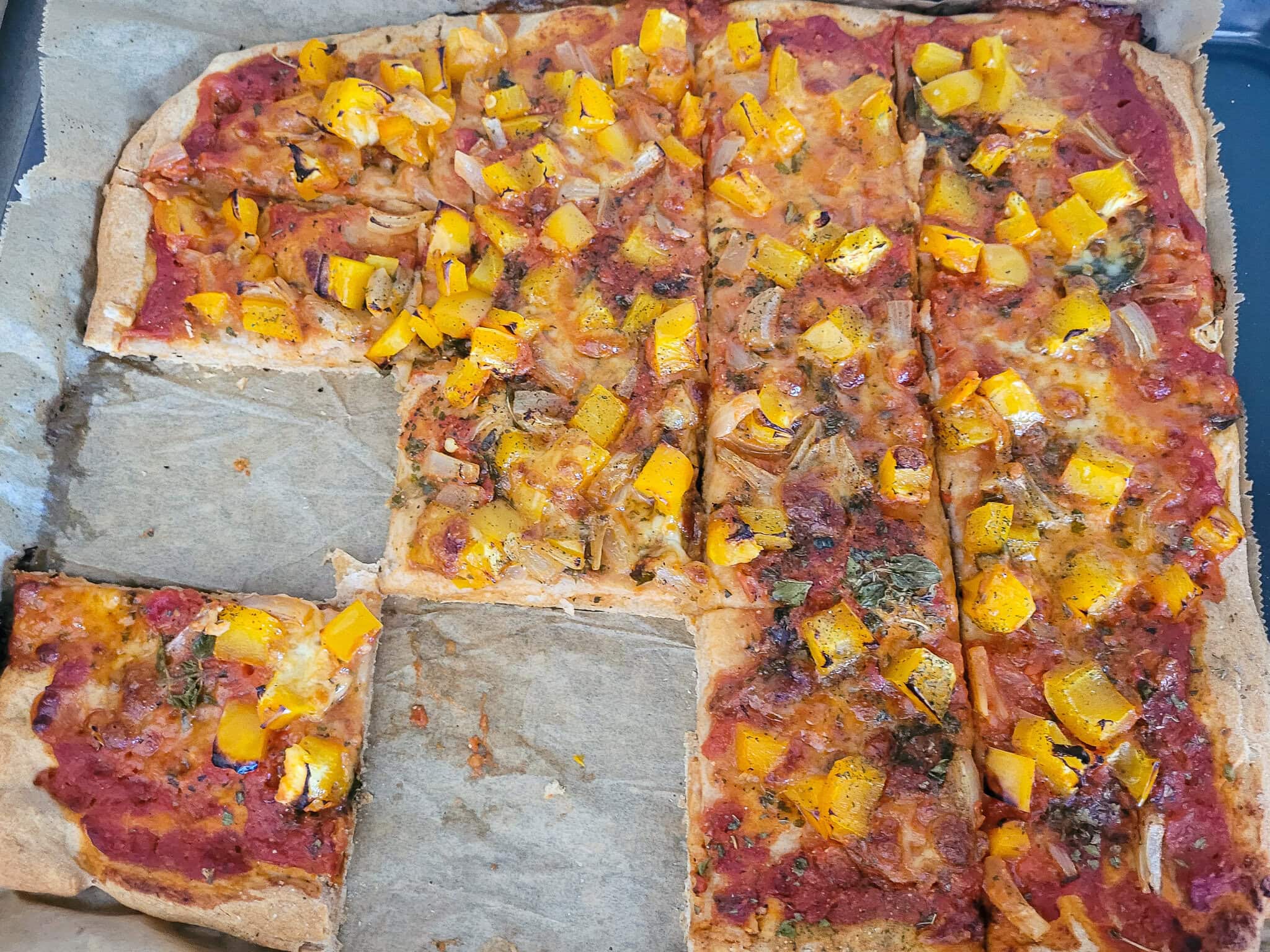 A close-up of a freshly baked bread dough pizza topped with yellow bell peppers, cheese, and other ingredients, on parchment paper.
