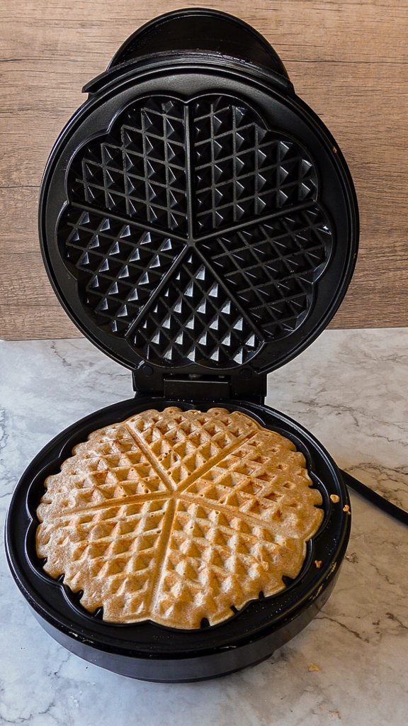 Black waffle maker with a cooked whole grain waffle inside, on a marble surface.