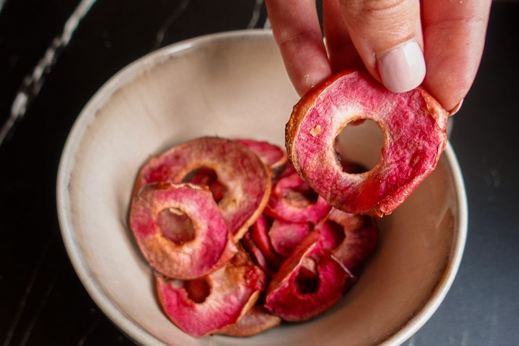 Hand holding a dried apple chip with a bowl of apple chips.