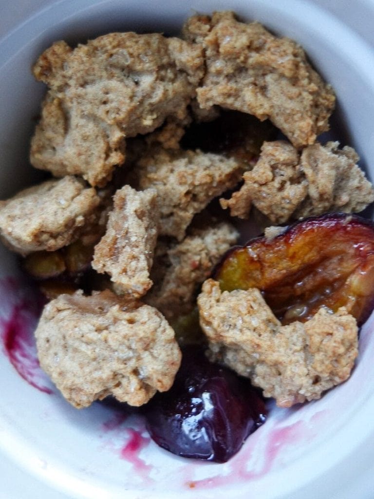 Close-up of a plum crumble in a white bowl with fresh plums and crumble topping.