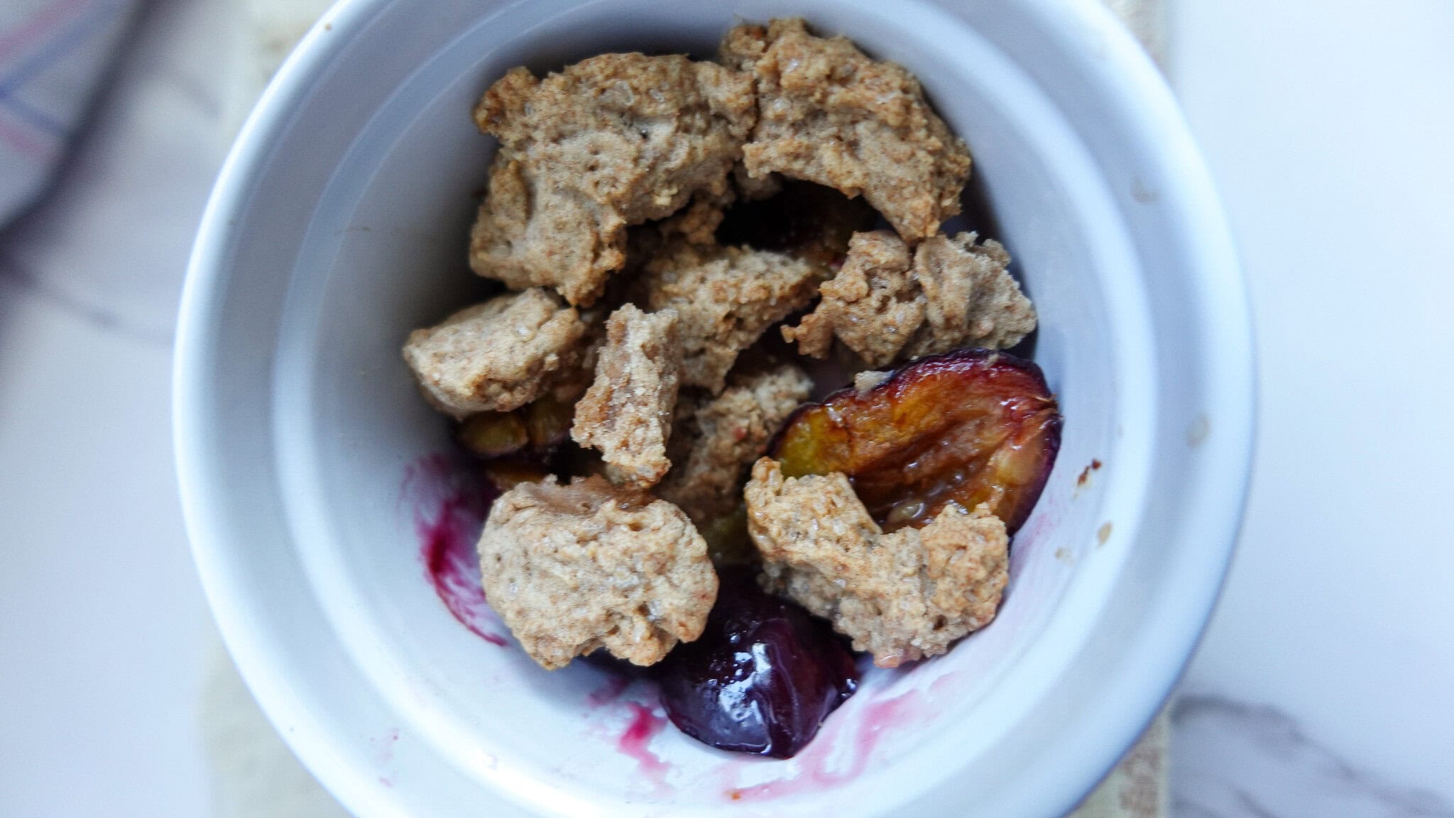 Close-up of a plum crumble in a white bowl with fresh plums and crumble topping.