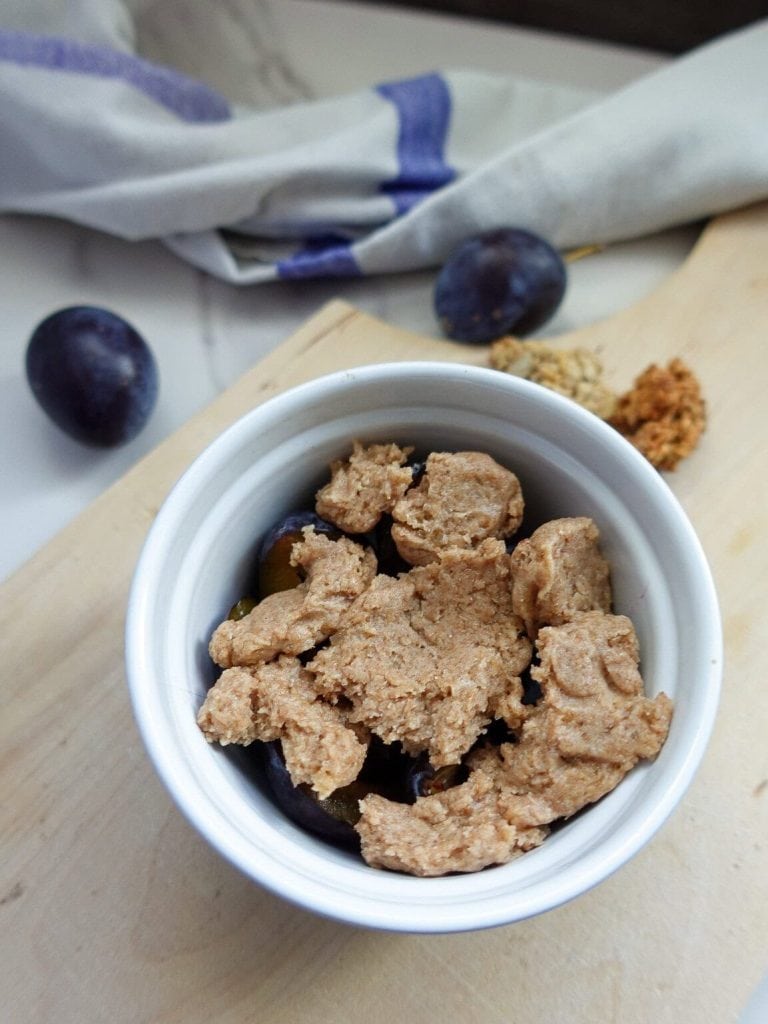 Close-up of plum crumble in a white bowl with fresh plums nearby.